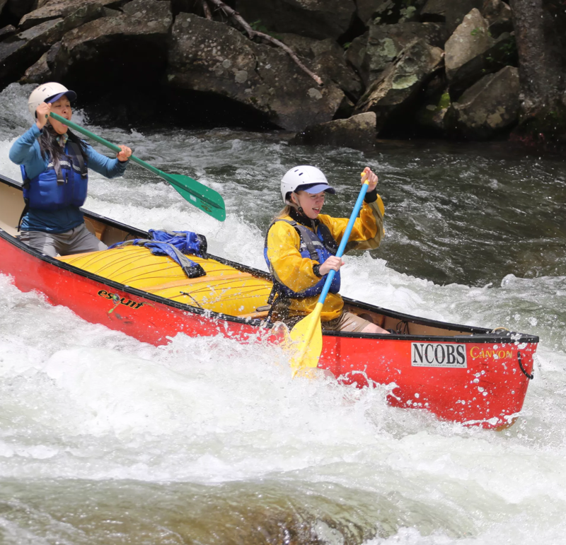 Two people are paddling a red and yellow canoe through some rapids. They are both wearing helmets and life jackets. The canoe has the letters 'NCOBS' on the side. The water is white and foamy, and there are rocks on either side of the river. The overall impression is one of adventure and excitement.